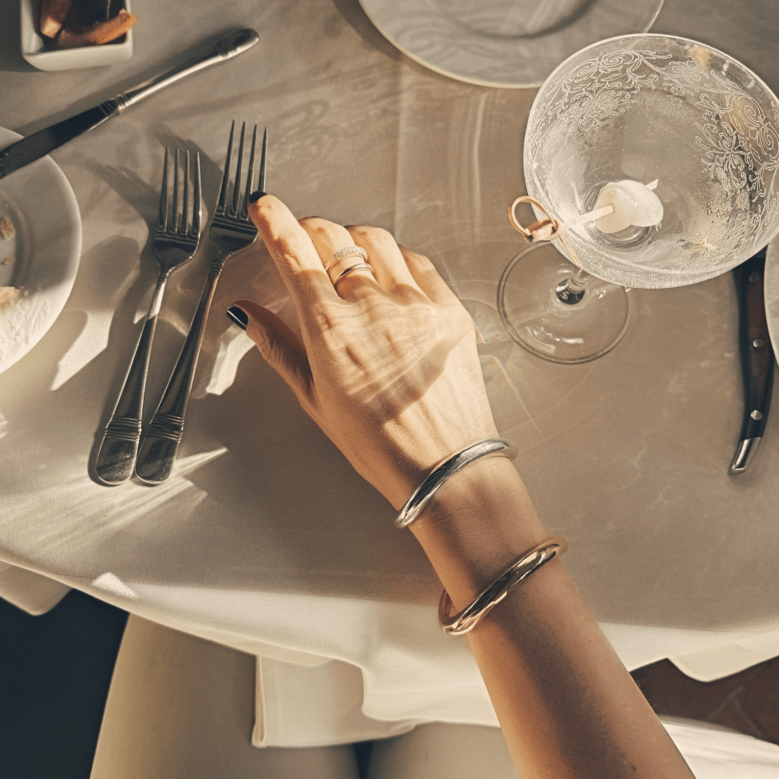 Person's hand with a ring on a table setting with cutlery and a glass and Gabriela Artiwas Wave Cuffs as a set