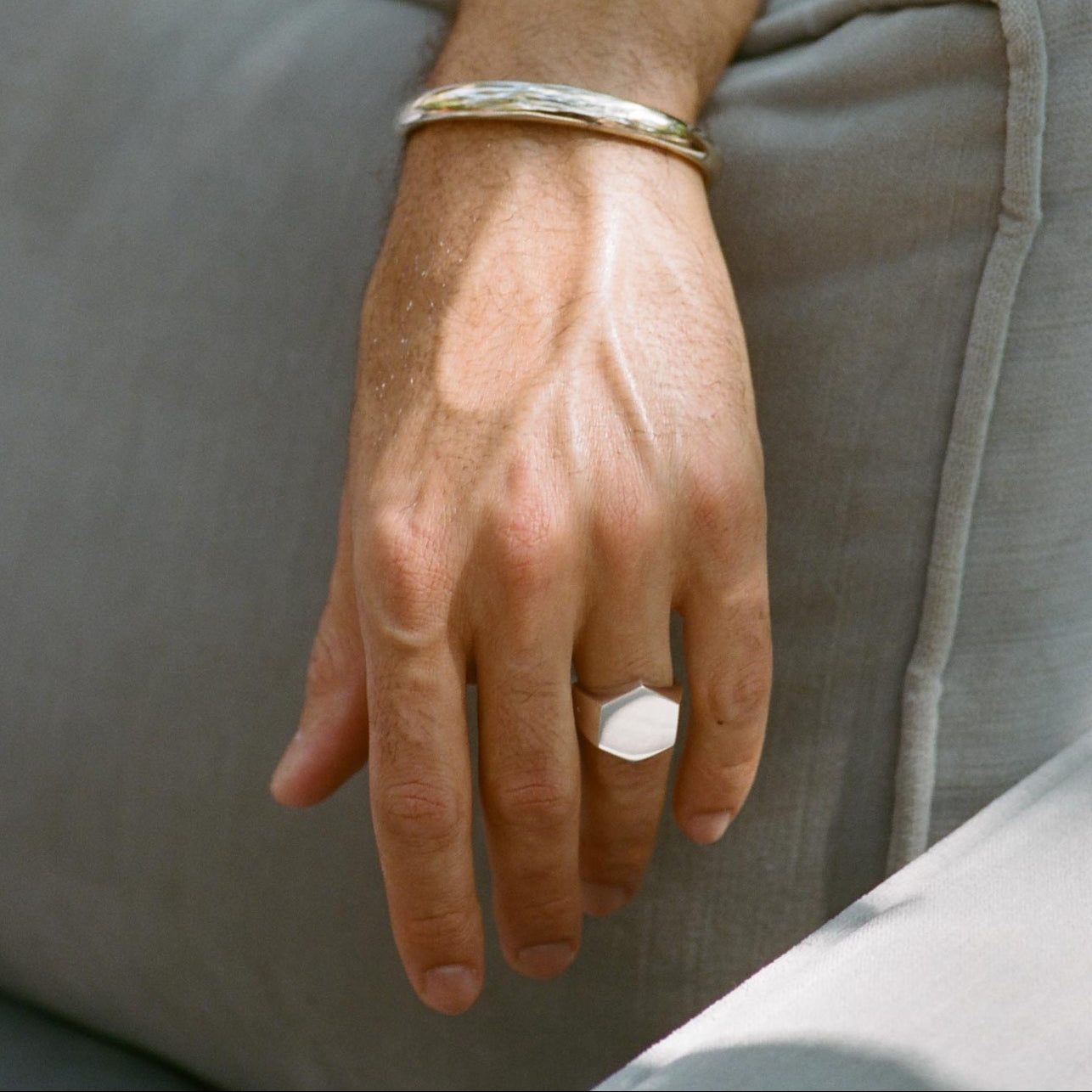Hand wearing a gold bracelet and silver flat hexigonal ring on a neutral background