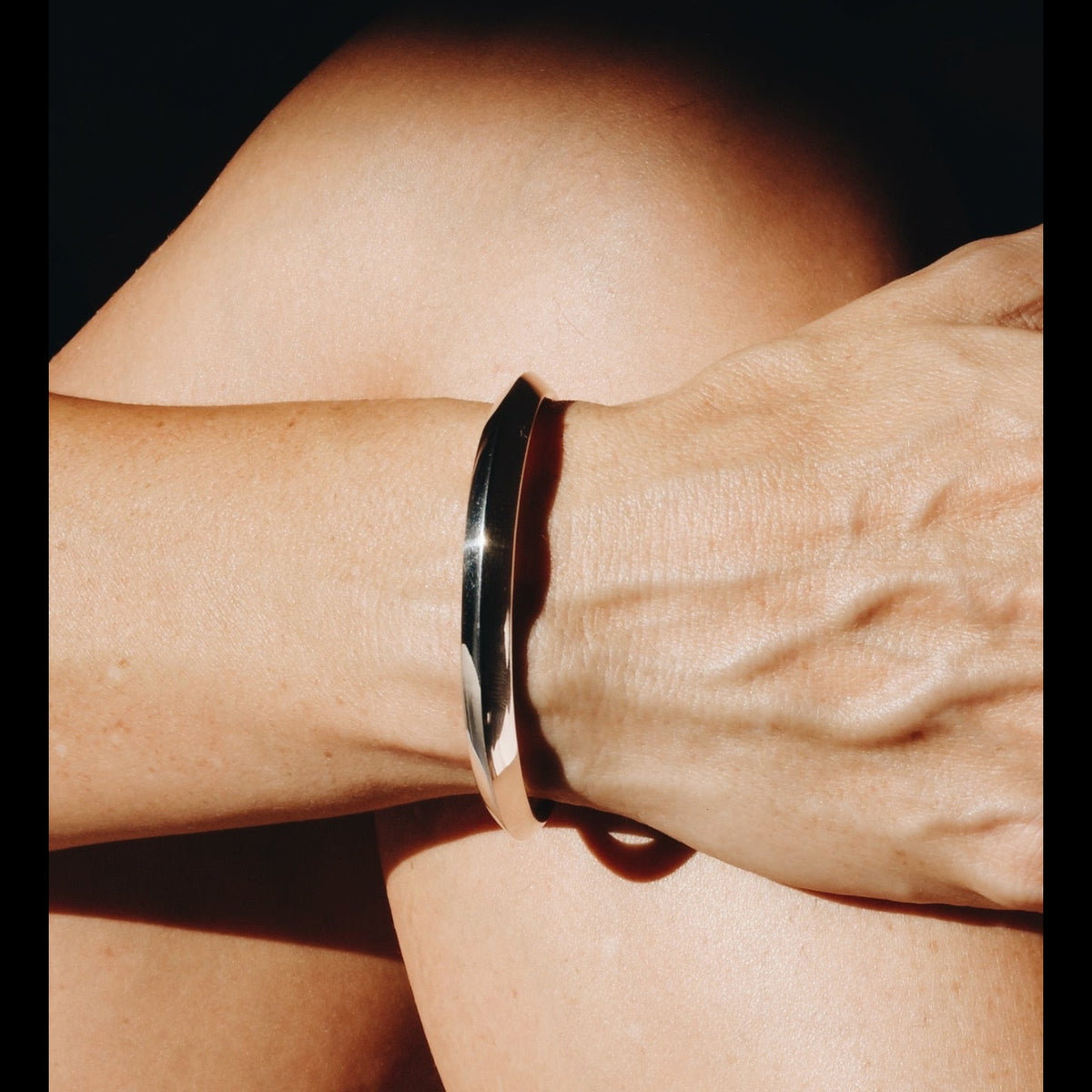 A close-up of a person's wrist wearing Gabriela Artigas Fang cuff in polished sterling silver metal cuff bracelet, with warm sunlight casting strong shadows against a dark background.